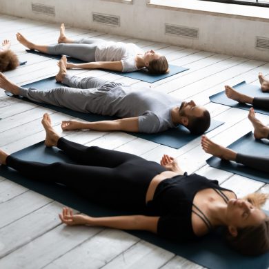 Young sporty people with closed eyes meditating in Savasana pose on floor, practicing yoga at group lesson, doing Corpse exercise on mats, training, working out in modern yoga studio, center